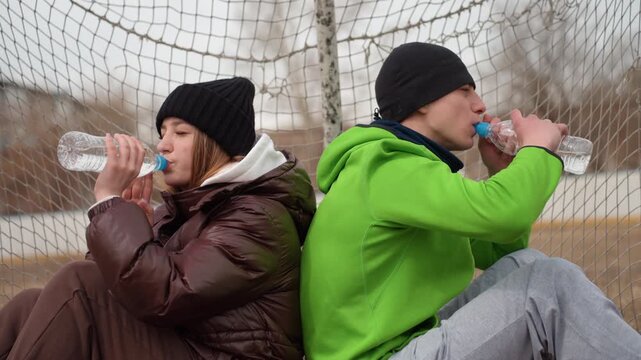 athletes hydrate warmly, two individuals sit back to back at sports goal while drinking water, man and woman of caucasian descent rest with drinks after intense winter exercise session