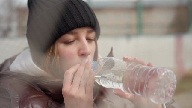 white woman drinking from plastic bottle beside goal net, black beanie and brown coat, chugging water for relief, companion in bright jacket behind, candid cold day recharge scene.