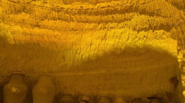 Ancient earthenware storage jars revealed as camera moves through a warm ochre chamber, showing handmade pottery arranged along a stone shelf in an archaeological interior