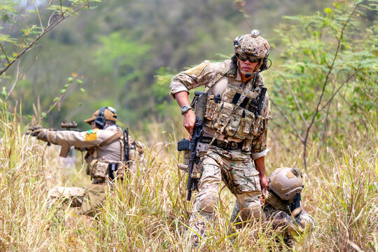 Three soldiers in full tactical gear advancing through tall grassy terrain with rifles ready, sunlight filtering through trees