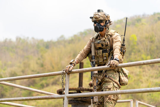 Soldier in full tactical gear leans on metal railing overlooking hilly jungle terrain with focused gaze
