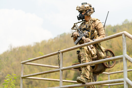 Soldier in full tactical gear leans on metal railing overlooking hilly jungle terrain with focused gaze
