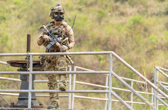 Soldier in full tactical gear stands alert on metal railing overlooking grassy hillside with rifle ready