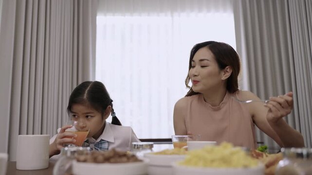 Happy Asian family mother and child daughter in school uniform sitting smiling while eating breakfast and drinking orange juice for health together before school during morning in the kitchen at home.