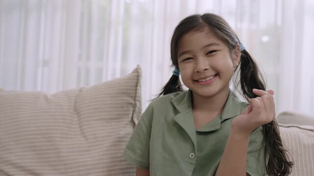 Portrait of happy cute Asian child girl sitting smiling with a mini heart and looking at camera on sofa in the living room at home. Little girl relaxing and cheerful on couch during vacation weekend. 