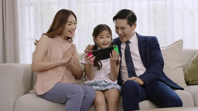 Happy Asian family father and mother sitting looking and cheering on their daughter while playing a game console, having fun and cheerful together, and she winner on sofa in the living room at home. 