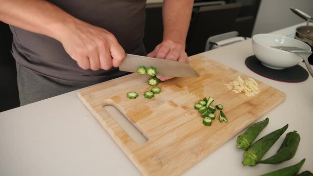 Male hands using a knife to chop okra and garlic on a wooden board in a modern kitchen