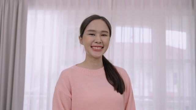Happy, confident, beautiful young Asian woman in pink clothes standing smiling and looking at the camera in the living room at home.  Portrait of a pretty Thai female model in a studio.