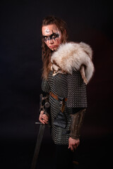 Obraz premium A young Viking woman in fur-trimmed chainmail and war paint, holding a sword, poses against a dark studio background.