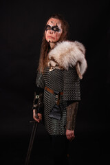 Obraz premium A young Viking woman in fur-trimmed chainmail and war paint, holding a sword, poses against a dark studio background.