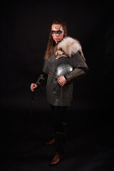 Obraz premium A young Viking woman in fur-trimmed chainmail and war paint, holding a sword, poses against a dark studio background.