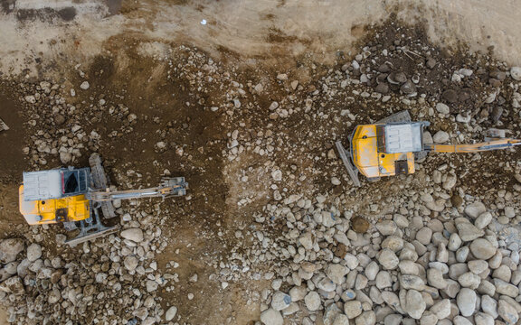 Yellow Excavators and a Heavy Truck Working Near Large Piles of Boulders and Rocks in a Rock and Sand Quarry in Indonesia. Mining Site and Heavy Machinery Viewed from Above.