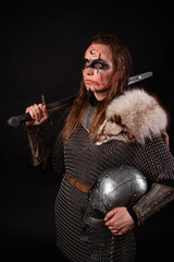 Obraz premium A young Viking woman in fur-trimmed chainmail and war paint, holding a sword, poses against a dark studio background.