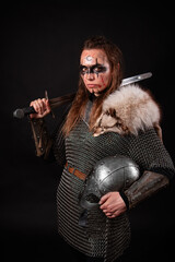 Obraz premium A young Viking woman in fur-trimmed chainmail and war paint, holding a sword, poses against a dark studio background.