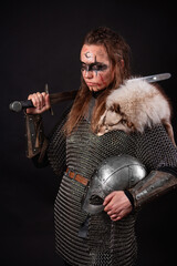 Obraz premium A young Viking woman in fur-trimmed chainmail and war paint, holding a sword, poses against a dark studio background.