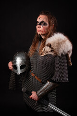 Obraz premium A young Viking woman in fur-trimmed chainmail and war paint, holding a sword, poses against a dark studio background.