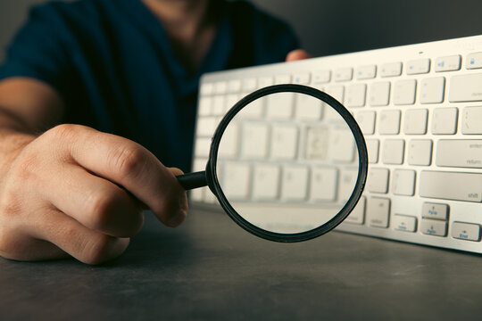 Man with magnifying glass and keyboard on grey background, closeup