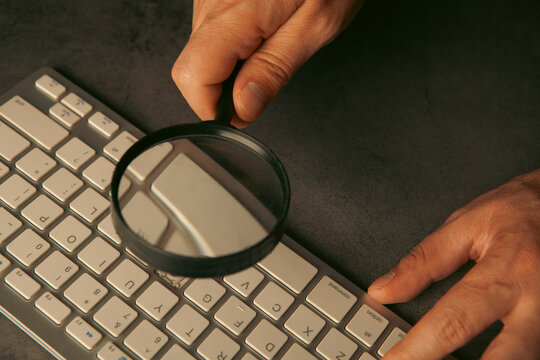 Man with magnifying glass and keyboard on grey background, closeup