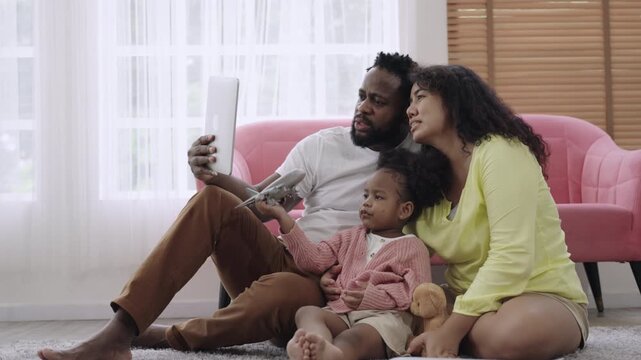 Happy Family Day concept. African American family sitting, smiling, and using a tablet for a video call while their little daughter plays with plane model, with happiness on living room floor at home.