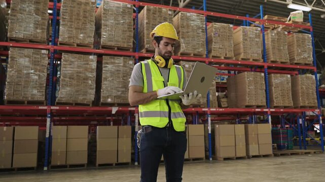 White man warehouse worker in uniform wearing a yellow safety helmet, standing, holding, and using a laptop computer for analysis while checking inventory stock in a logistics distribution factory. 