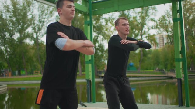 Two white men pausing at lakeside pavilion, breath catch and calm reflection, crossed arms, gaze over water, relaxed posture, supportive friendship, black training attire, serene park atmosphere