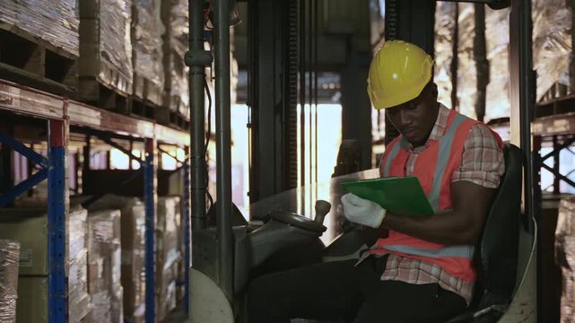 Professional African American male forklift driver wearing orange vest and yellow safety helmet, checking clipboard to inventory stock on shelves before delivery in a distribution warehouse center. 