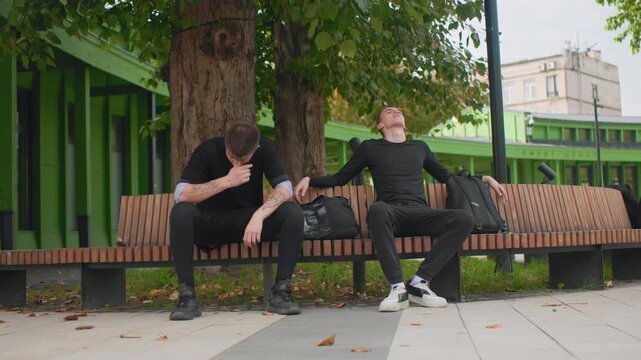 two white men sitting on bench under large tree near green pavilion, one slumps exhausted while companion scrolls phone, casual black clothing, backpacks on bench, autumn leaves on sidewalk, quiet