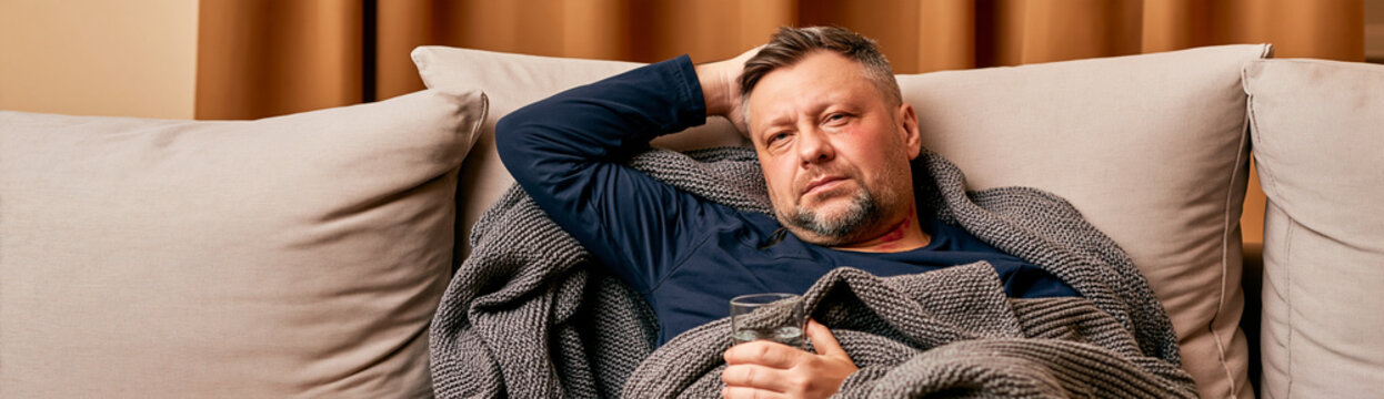 Middle-aged Caucasian man with flushed face, feverish and unwell, resting on sofa covered with blanket, holding glass of water, looking tired, conveying illness and fatigue at home