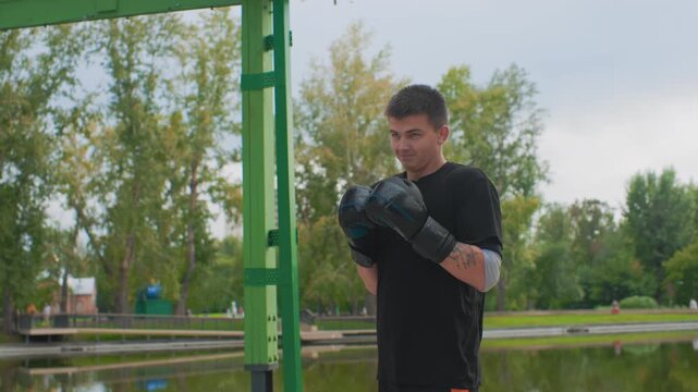 white man practicing shadowboxing with gloves by pond, adopting ready stance under green pergola, amateur athlete rehearsing technique, confident gaze, light breeze, urban park training
