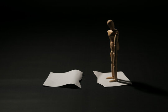Sad wooden mannequin standing on a torn piece of white paper looking down. Concept of separation, divorce, broken contract, failure, or relationship gap.