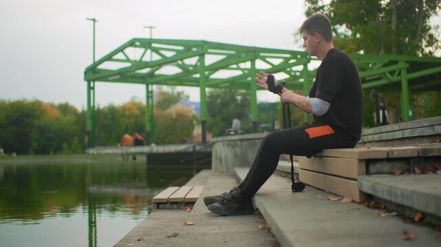 young man checking phone by pond bench, scrolling messages and adjusting camera, seated on concrete steps near green bridge, calm water reflections and casual autumn park atmosphere
