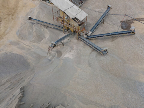 Aerial View of Rock Crusher Machinery and a Conveyor Belt System Near Large Aggregate Stockpiles at an Industrial Quarry Site. Rock Crushing and Aggregate Production Viewed from Above.