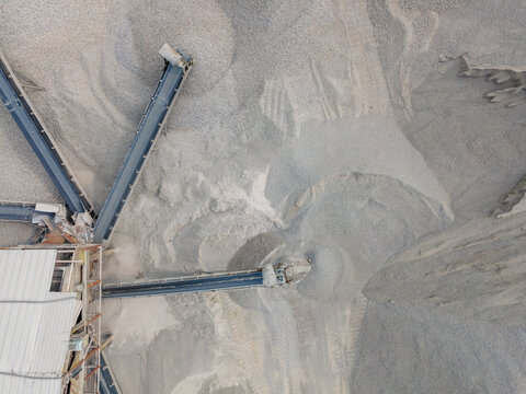 Aerial View of Rock Crusher Machinery and a Conveyor Belt System Near Large Aggregate Stockpiles at an Industrial Quarry Site. Rock Crushing and Aggregate Production Viewed from Above.
