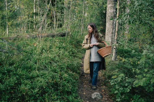 Woman walking along a narrow woodland trail with wicker basket while searching the forest floor, illustrating seasonal foraging, mushroom picking and quiet nature exploration.