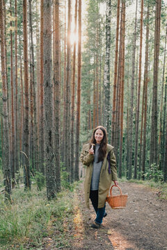 Woman walking through a sunlit pine forest with wicker basket and freshly picked mushroom, illustrating seasonal foraging, mushroom hunting and peaceful outdoor lifestyle.