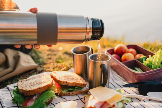 Hot drink being poured from a thermos into metal mugs during an outdoor picnic with sandwiches, fruit and vegetables beside a lake in warm golden light.