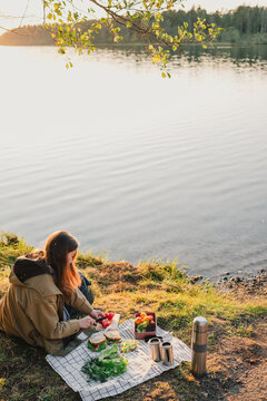 Woman preparing fresh sandwiches on a picnic blanket beside a calm lake at golden hour, enjoying healthy outdoor food, peaceful nature leisure and lakeside relaxation.