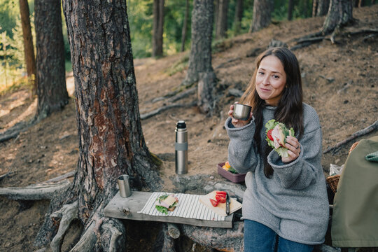 Woman enjoying a forest picnic with a fresh sandwich and hot drink, relaxing beside a pine tree with thermos, healthy food and peaceful outdoor lifestyle.