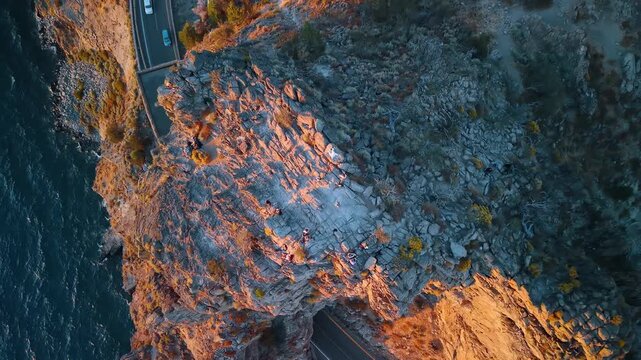 Overhead view of Cave Rock and the shoreline highway. Orange sunlight touches fractured stone beside a road, with dark blue water along the left edge.