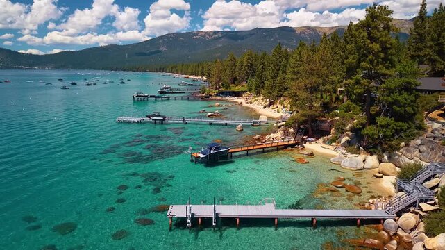 Piers and boats along the turquoise shore of Lake Tahoe. Docks extend from a conifer lined beach over submerged rocks, with mountains beneath scattered clouds.