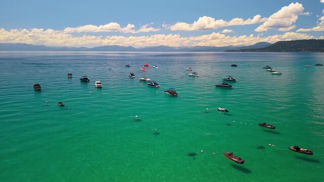 Moored boats across turquoise Lake Tahoe water. Small vessels and buoys dot the bay beneath a blue sky, with mountains and white clouds on the horizon.
