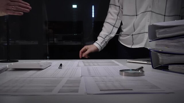 Business professionals shake hands over a cluttered desk of contracts, calculator and magnifying glass, sealing a trusted partnership after detailed financial review and late night work
