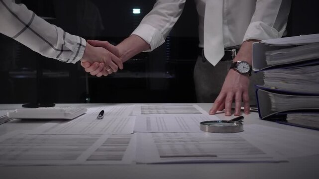 Business professionals shake hands over a cluttered desk of contracts, calculator and magnifying glass, sealing a trusted partnership after detailed financial review and late night work