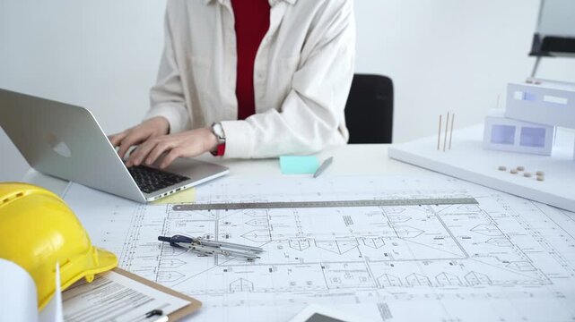 Architect working on new residential project using laptop, reviewing blueprints and drafting floor plans for modern building design with a 3d model on office desk