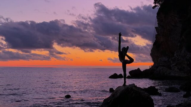 A woman silhouette holds a dancer yoga pose on a rock beside the sea at sunset, arm raised and one leg bent overhead, near a coastal cliff in Budva, a concept of balance and serenity.