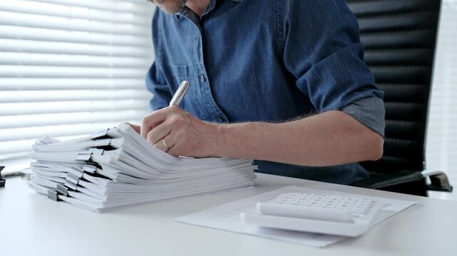 Individual working diligently on financial documents, calculating expenses and reviewing reports at an office desk, managing a large stack of paperwork and invoices