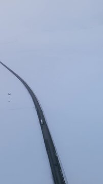 Drone view of a long asphalt road cutting through vast white snowfields at Ala-Bel Pass in the Tien Shan mountains, Kyrgyzstan, with cars driving in winter weather