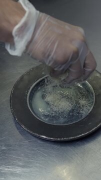 A gloved cook sprinkles fine shredded pastry into a greased round pan to form the base of a kunafa dessert.