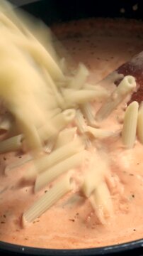 cook adds penne pasta to tomato garlic cream sauce in pan in home kitchen; process of preparing tomato based pasta dish; delicious and healthy Italian meal is being created; food closeup