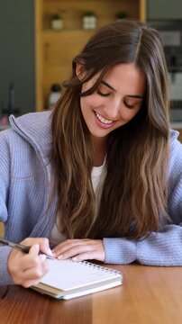 Beautiful smiling brunette woman sitting at a wooden table, writing down her thoughts, plans, and creative ideas in a notebook, feeling inspired and content in her cozy home environment